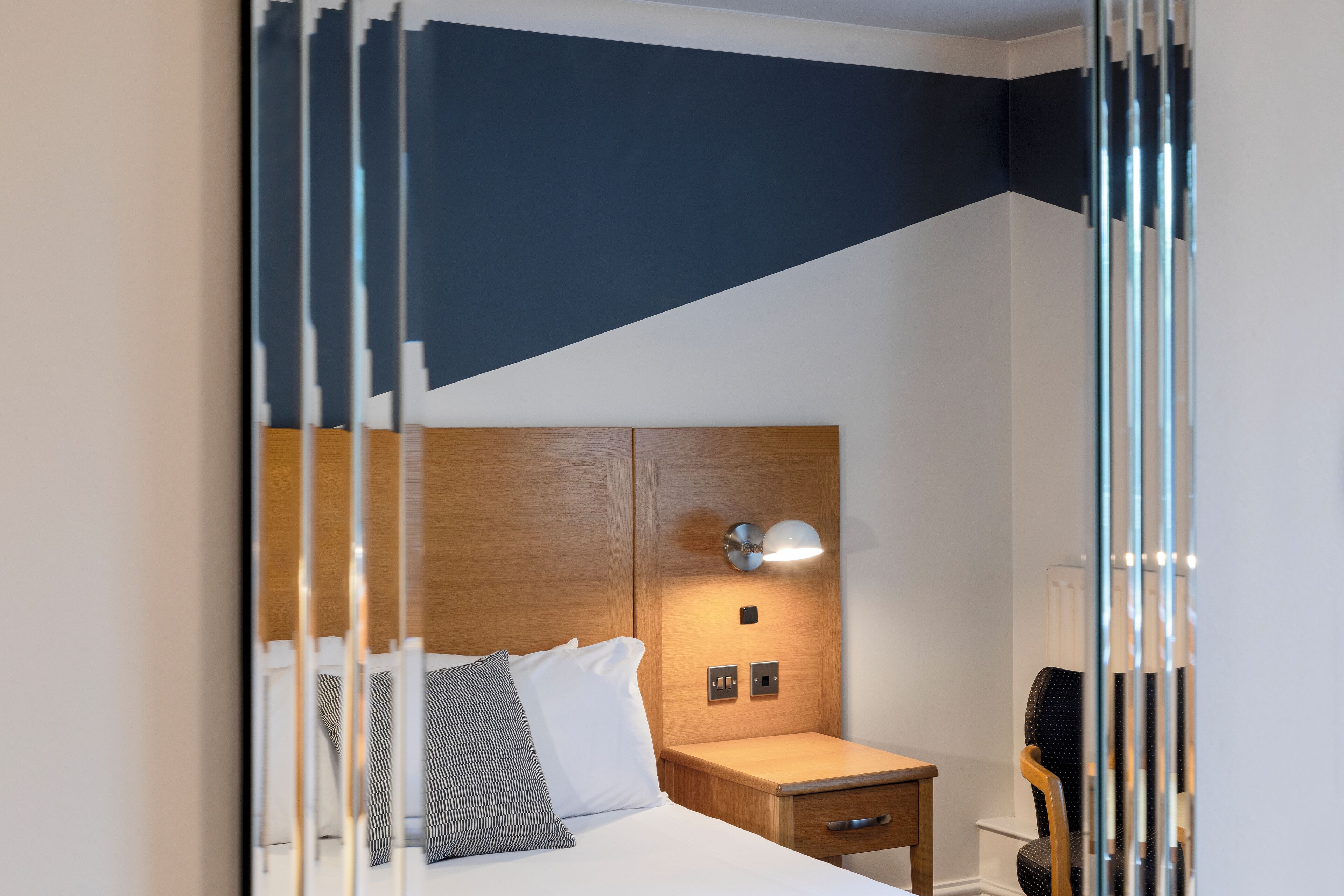 A modern hotel room with a wooden headboard, a bedside table with a lamp, and a chair.