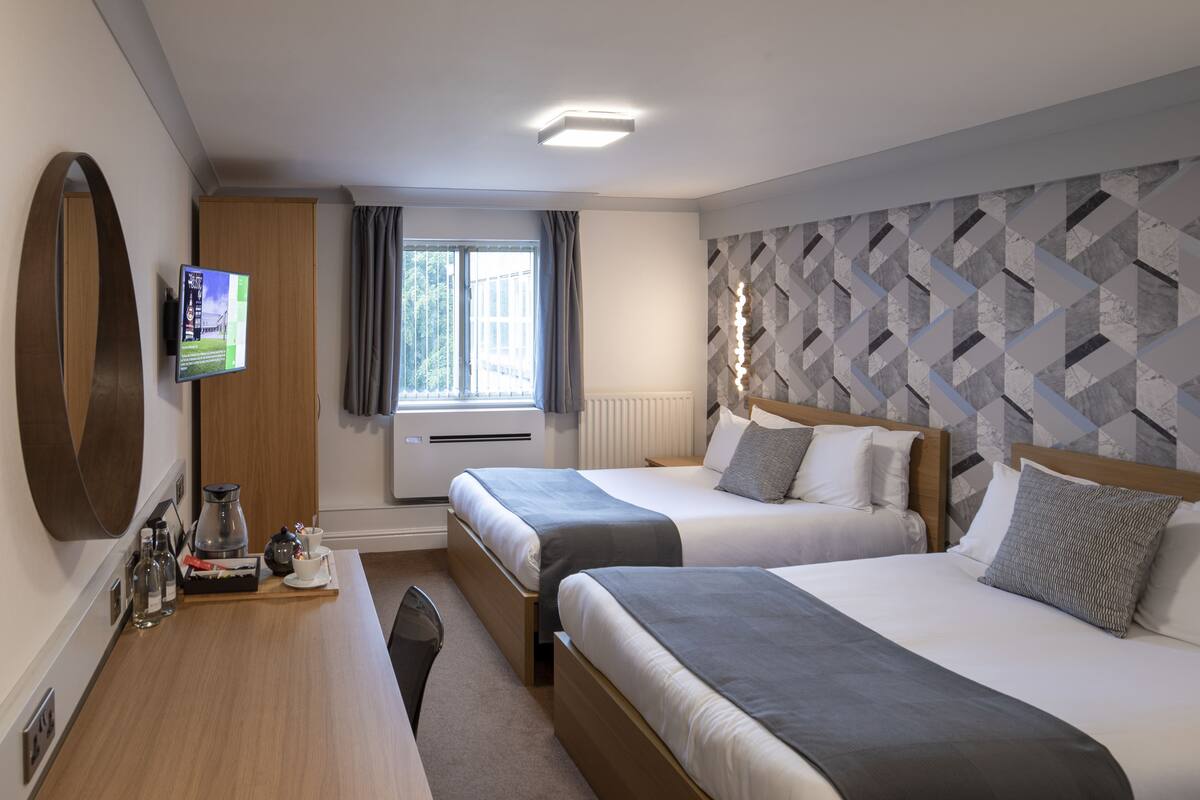 A hotel room with two beds, a TV mounted on the wall, a desk with a coffee maker, and a window with curtains.