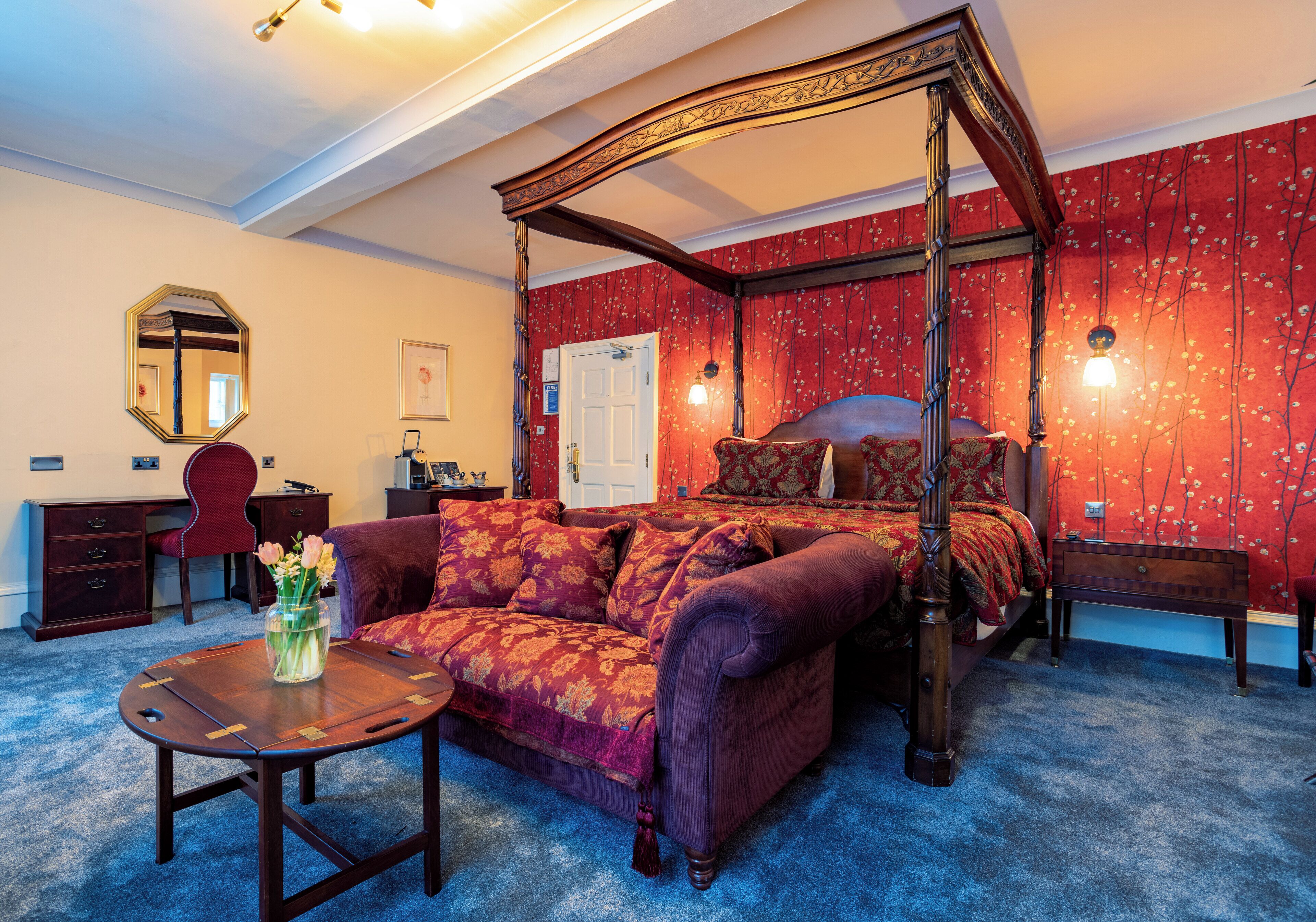 A four-poster bed with a canopy, a red floral wallpaper, a wooden desk, a red armchair, and a small table with a vase of flowers.