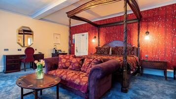 A four-poster bed with a canopy, a red floral wallpaper, a wooden desk, a red armchair, and a small table with a vase of flowers.