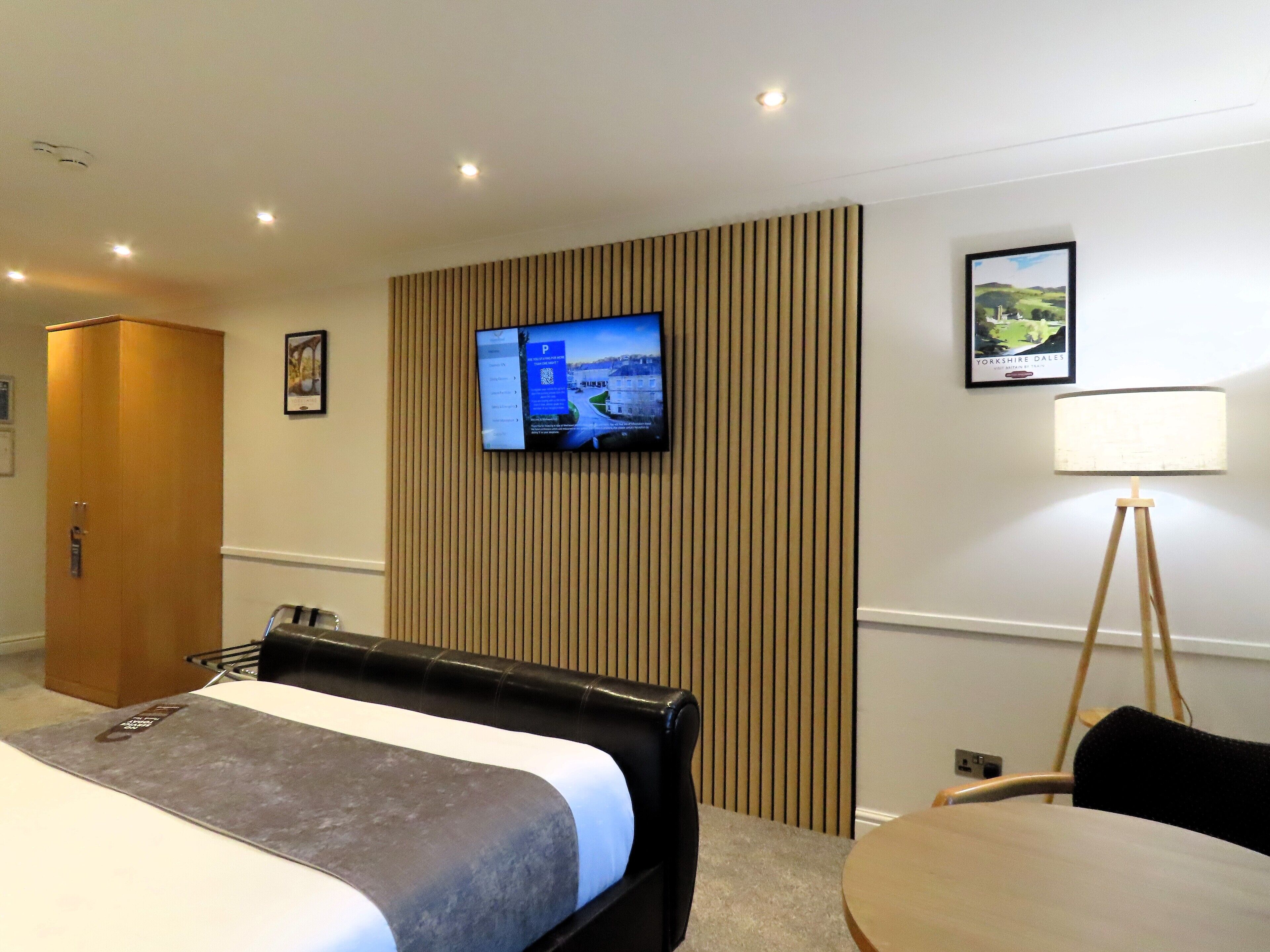 A hotel room with a bed, a TV mounted on a wall, a desk with a chair, and a lamp.
