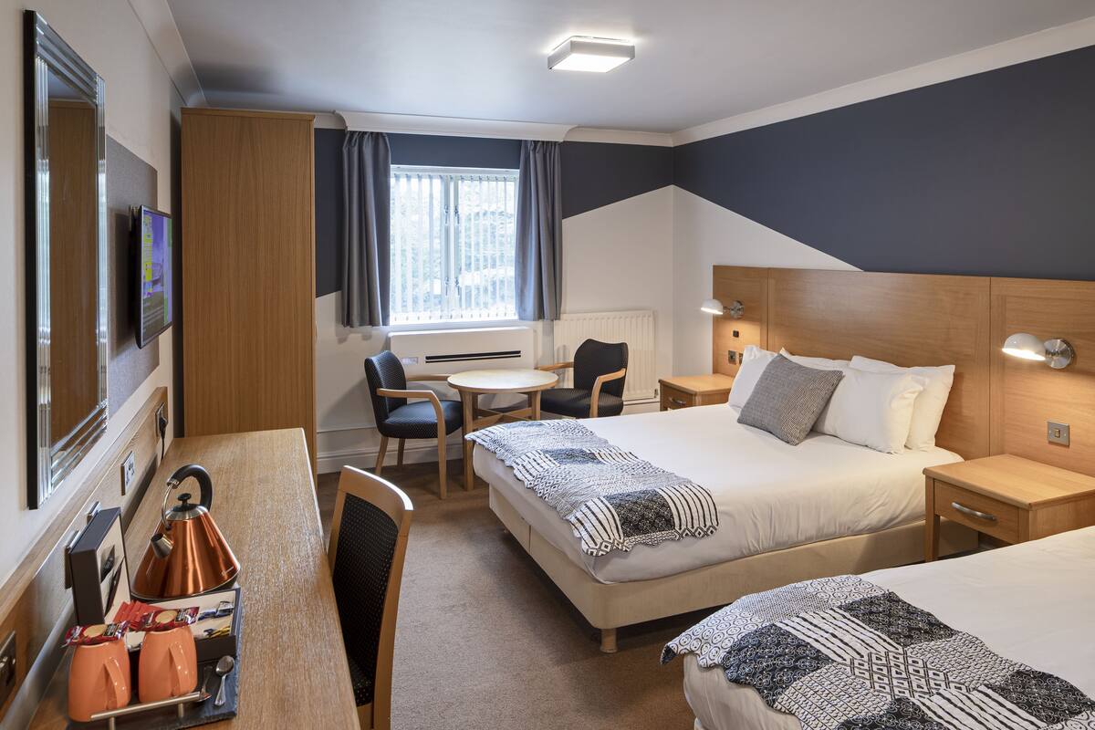 A hotel room with two beds, a desk with a kettle, a chair, a small table, and a TV.