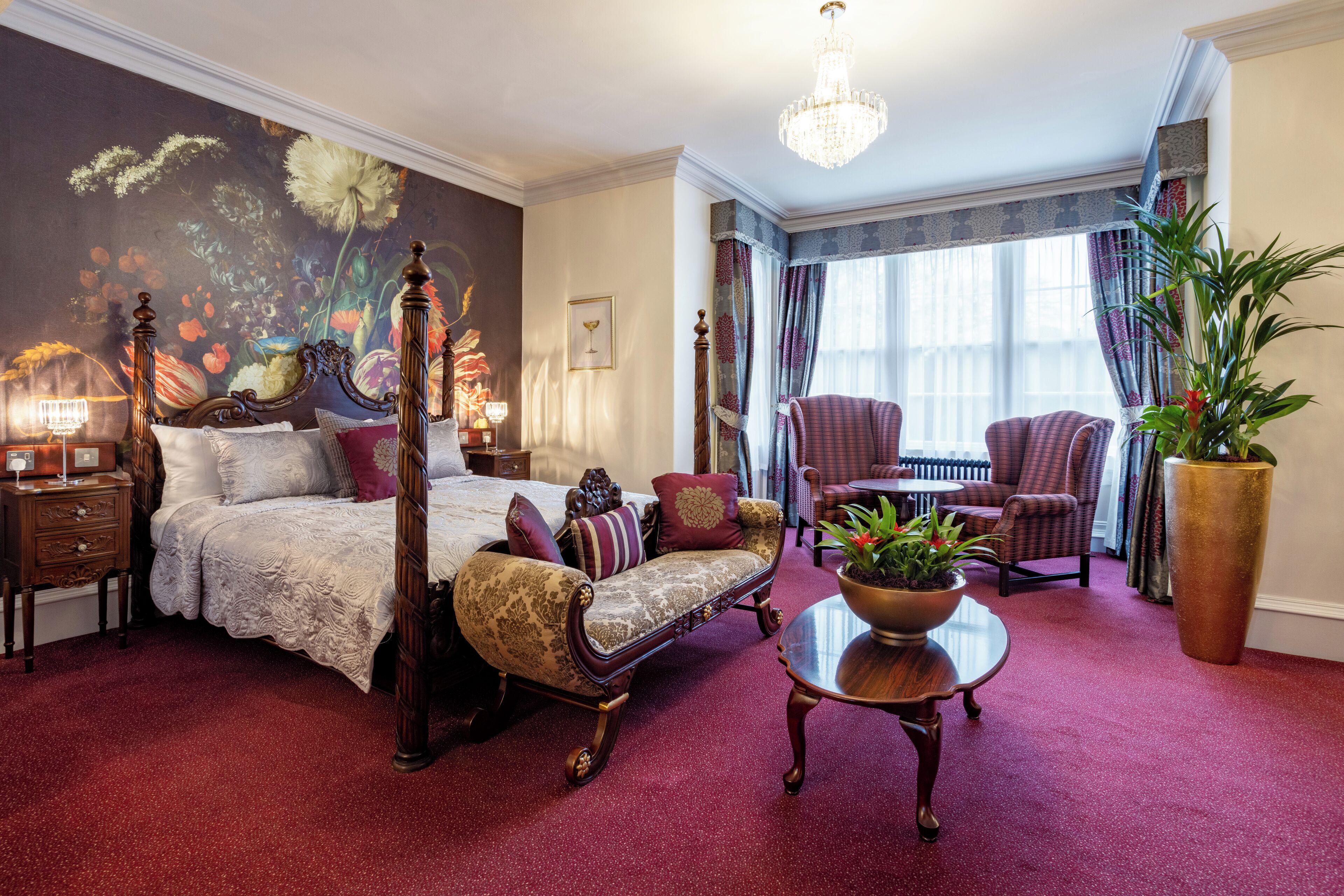 A bedroom with a four-poster bed, a sitting area with a chair and a table, and a large potted plant.