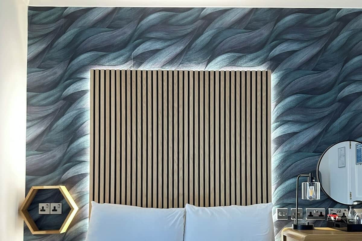 A modern hotel room with a large bed, a wooden headboard, a bedside table with a lamp, a mirror, and a hexagonal wall decoration.