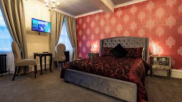 A bedroom with a large bed, a television mounted on the wall, a chair, a small table, and a nightstand with a lamp.