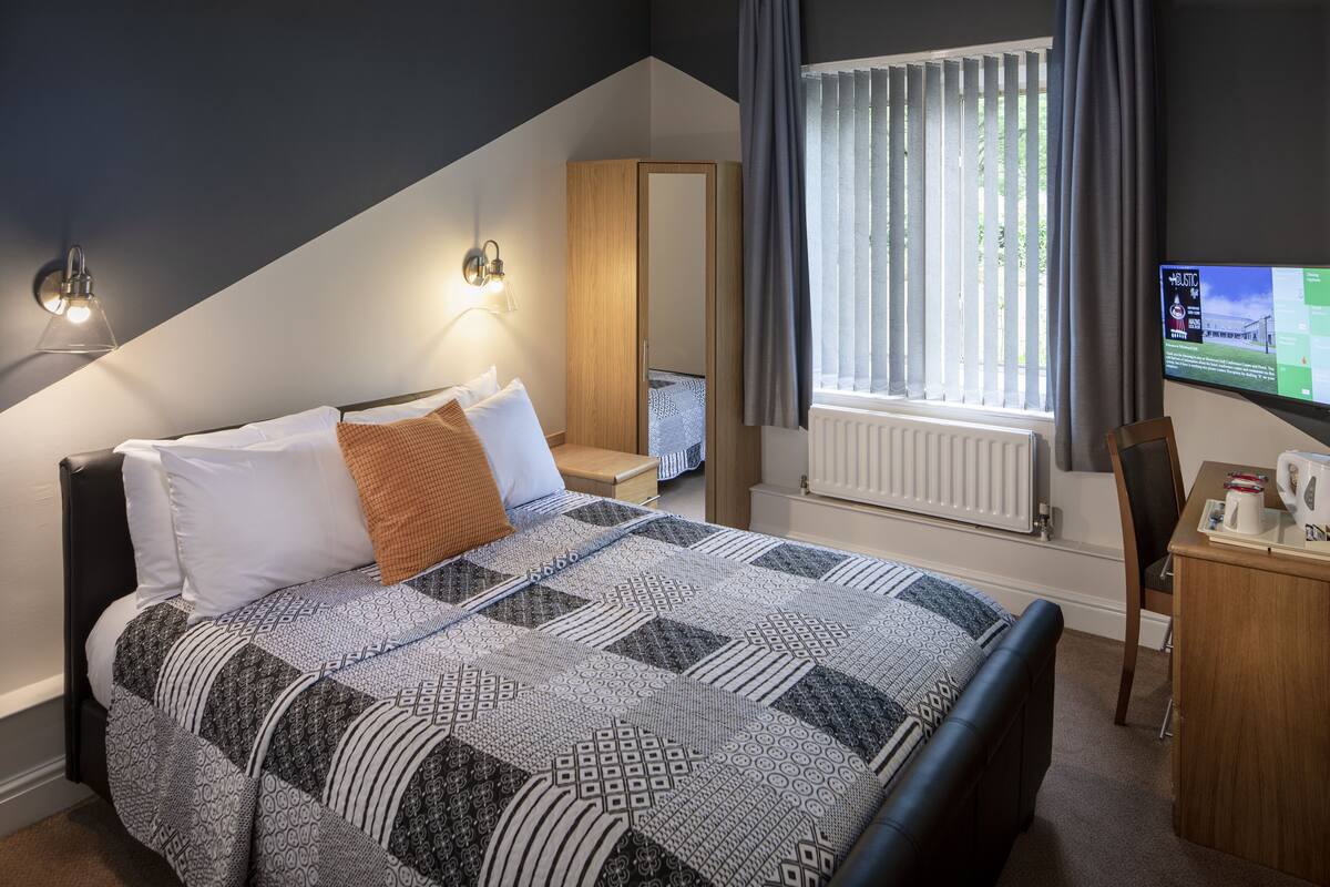 A bedroom with a bed, a desk, a chair, a TV, and a window with curtains.