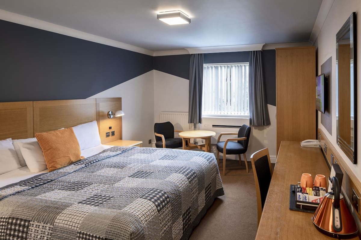 A hotel room with a large bed, a desk, two chairs, a small table, and a window with curtains.