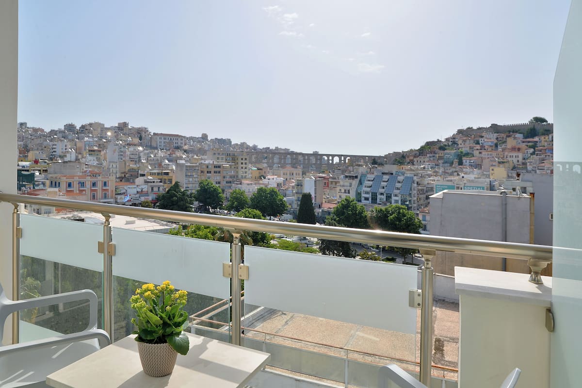 A balcony with a potted plant, a railing, and a view of a densely built cityscape with various buildings and a bridge in the distance.