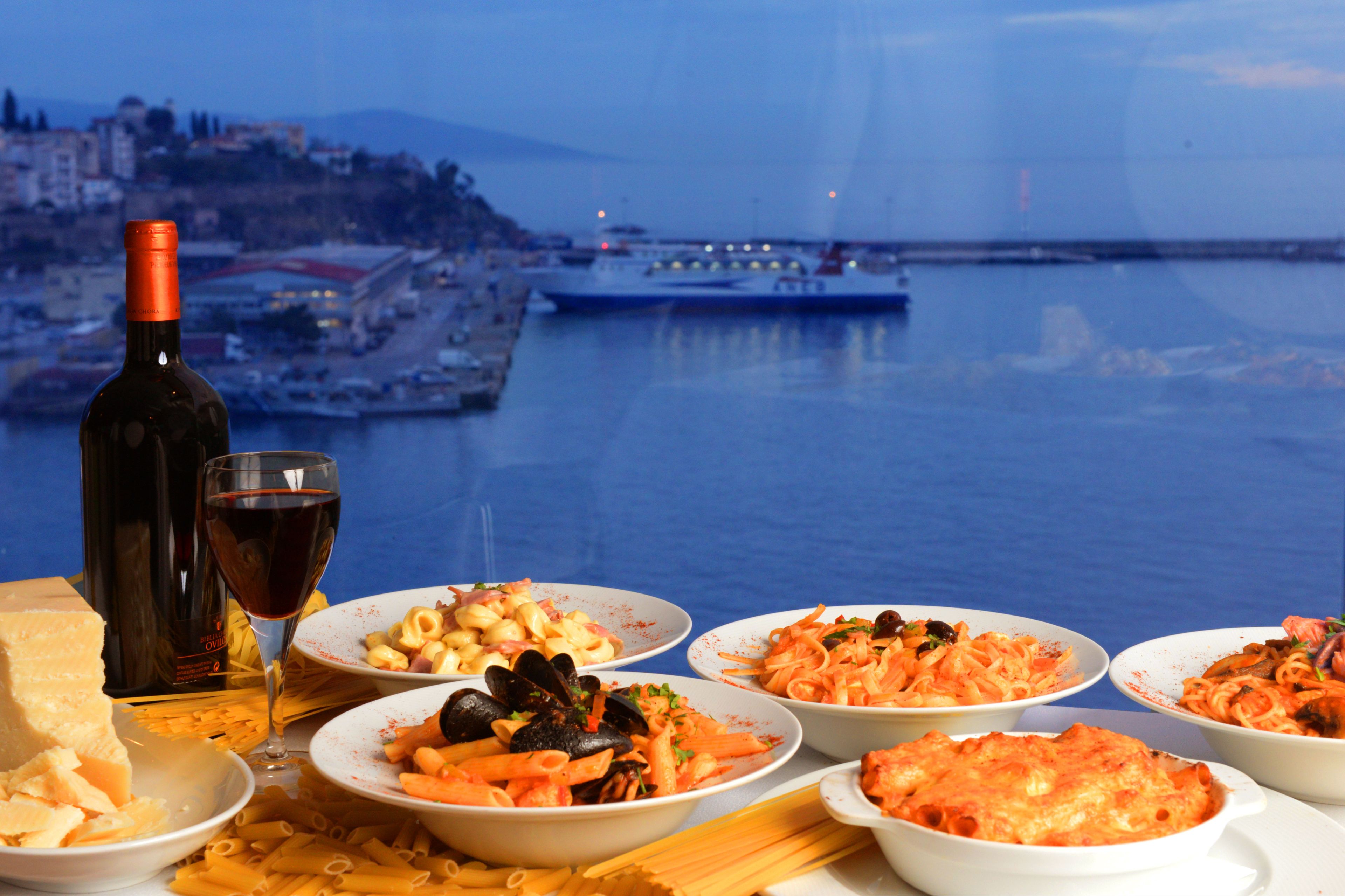Una mesa con varios platos y una botella de vino, con vista a un puerto con un barco y edificios al fondo.