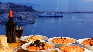 Una mesa con varios platos y una botella de vino, con vista a un puerto con un barco y edificios al fondo.