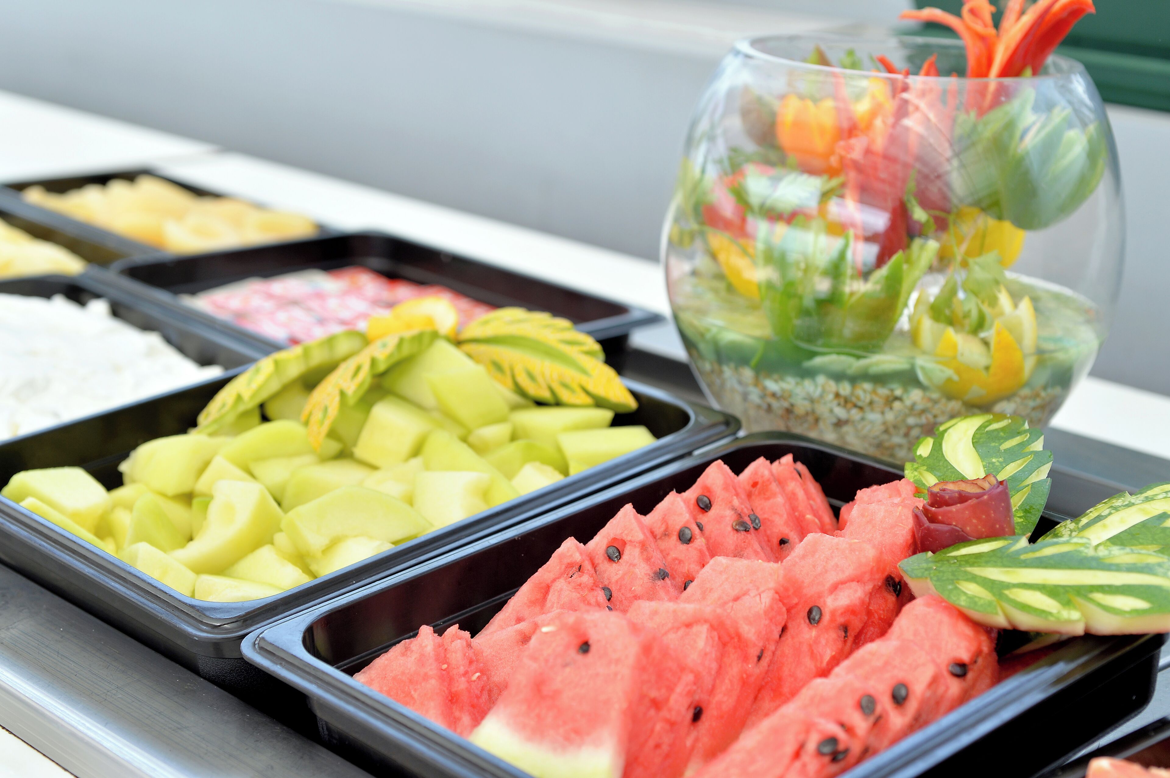 Un bufé con diversas frutas y verduras, incluyendo sandía, melón, y un cuenco de vidrio decorado con flores.