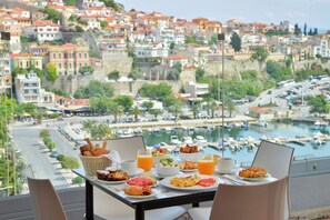 Una mesa con productos para el desayuno y un ordenador portátil, con vistas a un puerto deportivo y una ciudad en la colina.