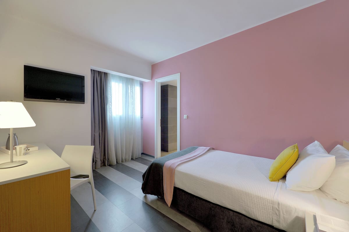 A hotel room with a bed, a desk with a lamp, a TV mounted on the wall, and a window with curtains.