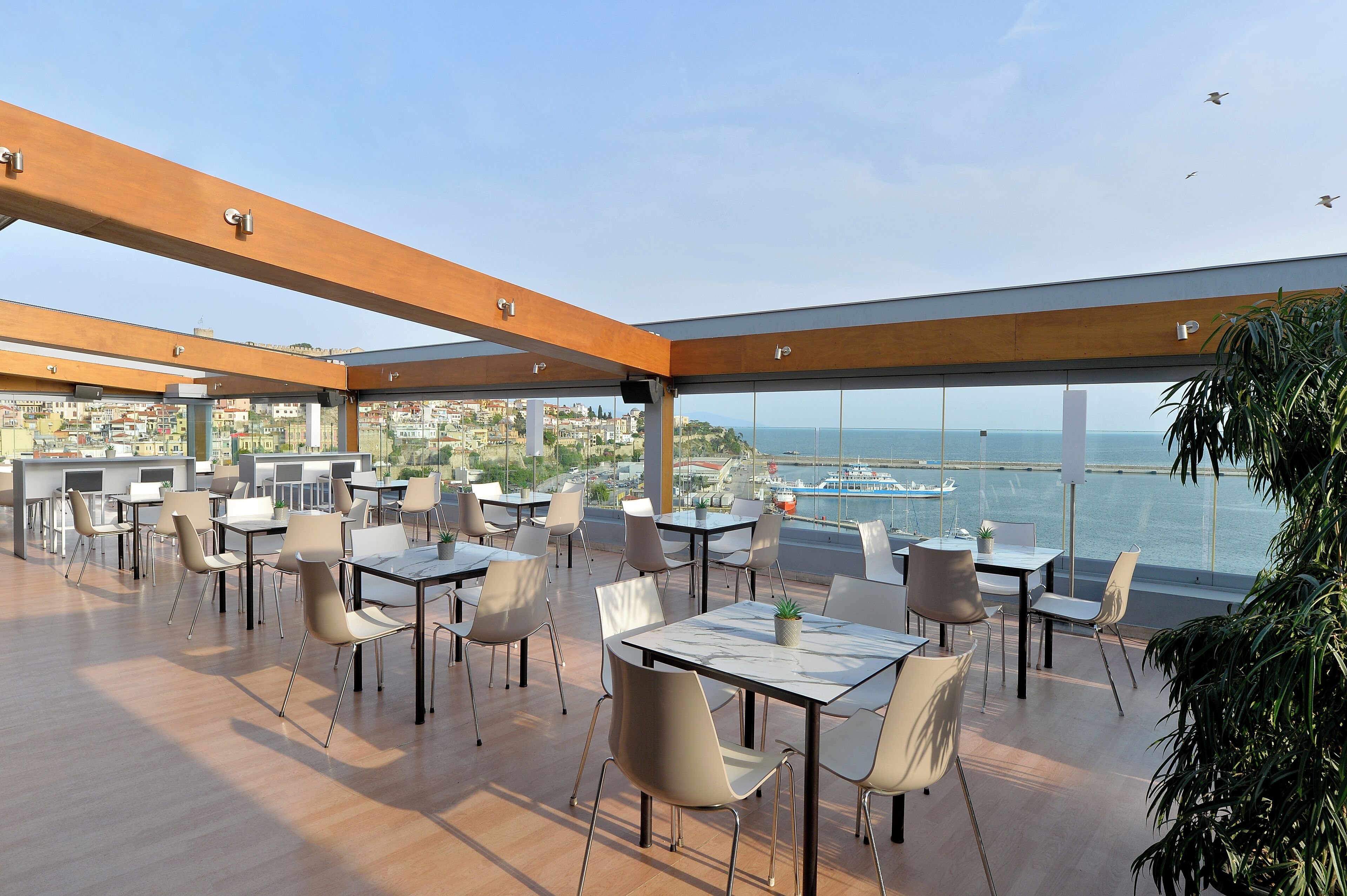 A rooftop dining area with tables and chairs, large windows, and a view of the sea and cityscape.