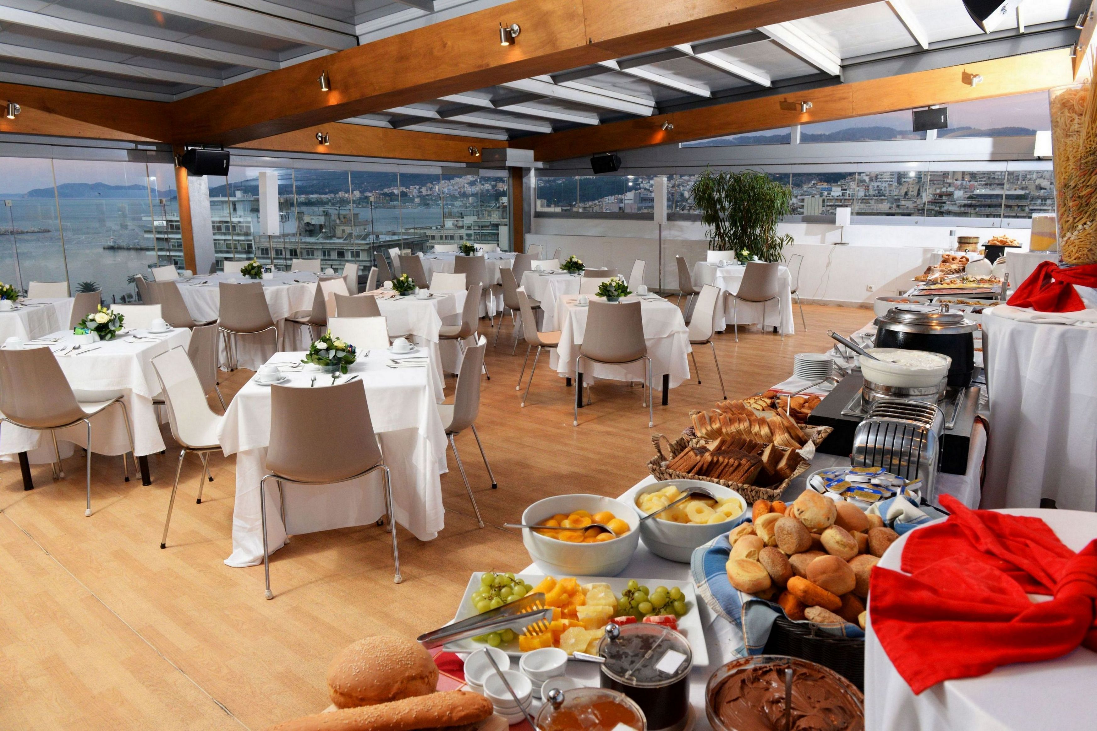 Un área de comedor en la azotea con mesas preparadas para una comida, un bufé con diversas opciones de alimentos y vistas a la ciudad.