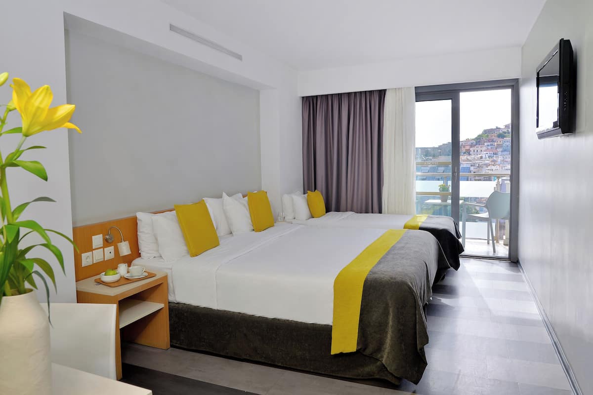 A hotel room with a large bed, a bedside table with a vase of flowers, a view of a cityscape through a large window, and a flat-screen TV mounted on the wall.
