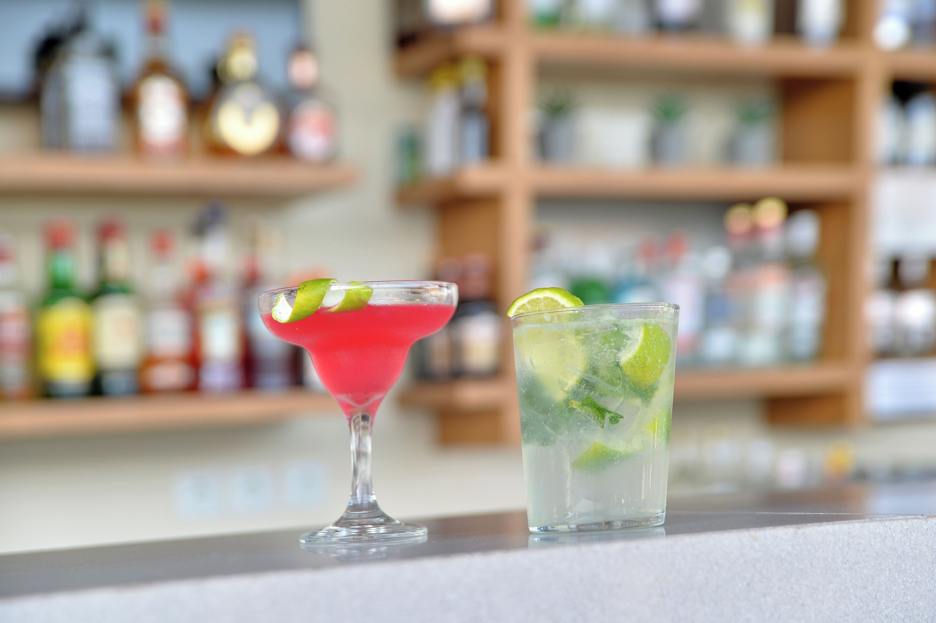 Two cocktails on a bar counter with shelves of bottles in the background.