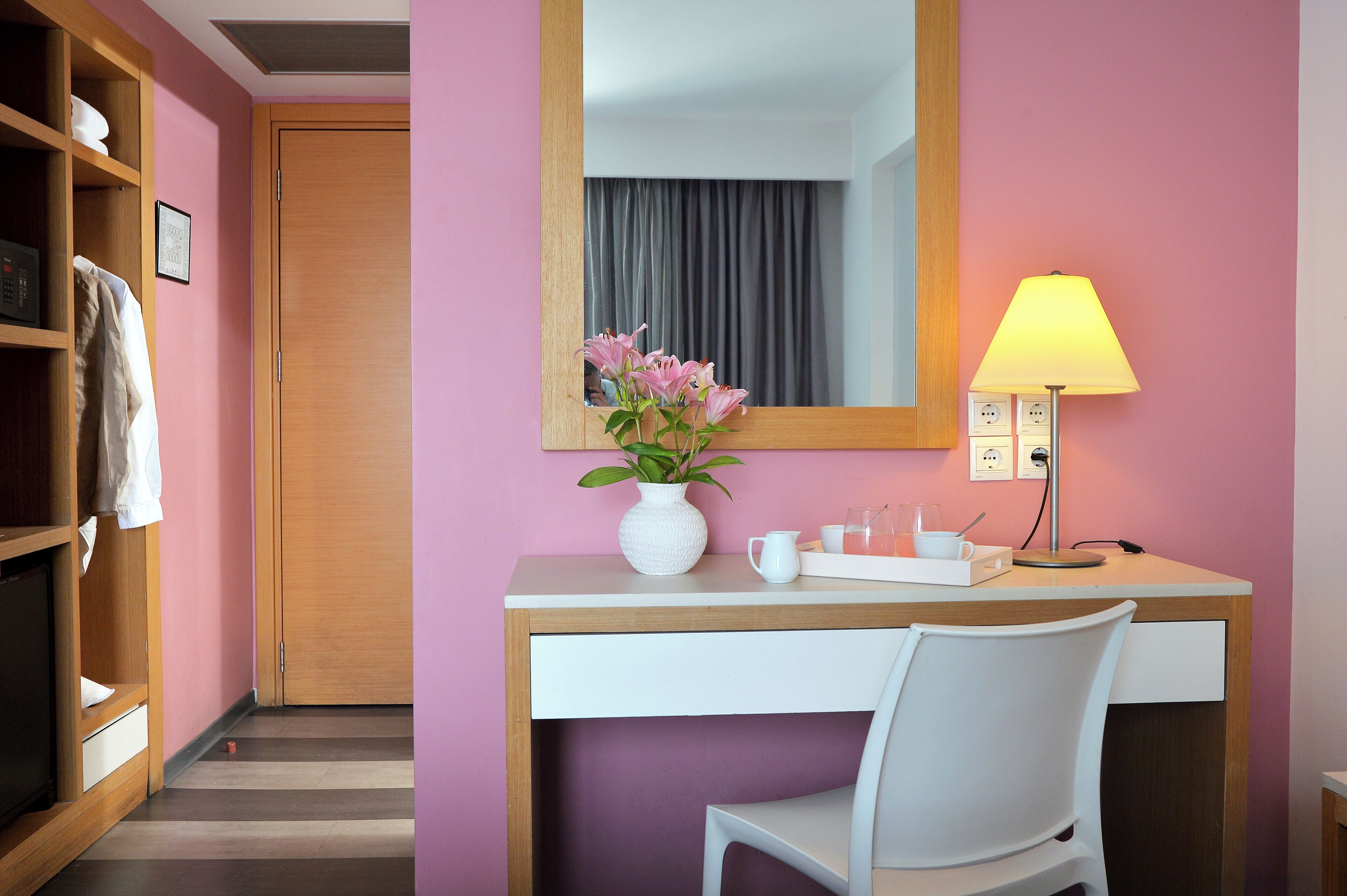 Una camera d'albergo con una scrivania in legno, una sedia bianca, un vaso con fiori, una lampada e uno specchio.