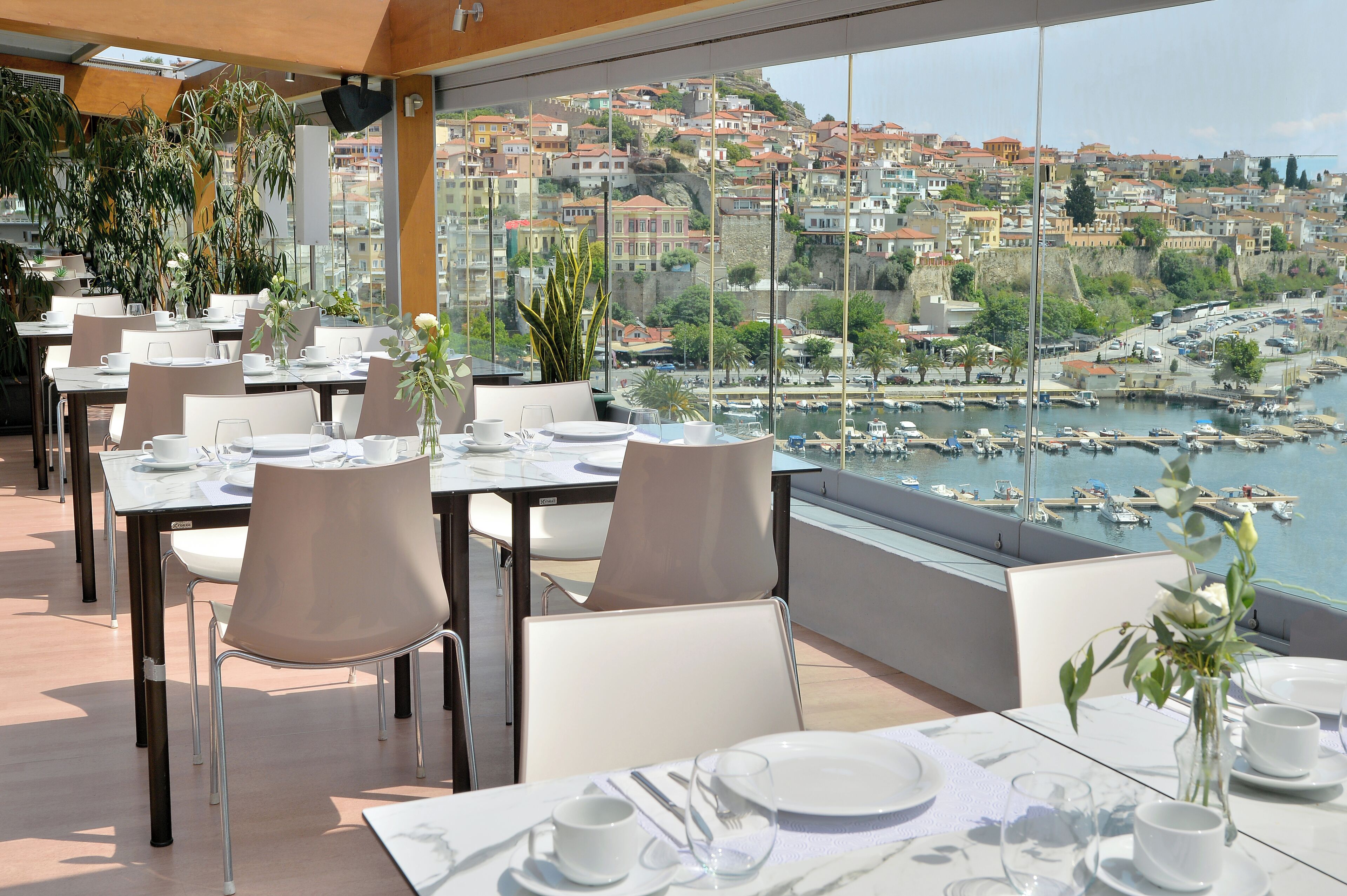 Un restaurante con mesas dispuestas para comer, con vistas a un puerto deportivo y una ciudad en la ladera.