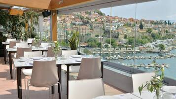 Un restaurante con mesas dispuestas para comer, con vistas a un puerto deportivo y una ciudad en la ladera.