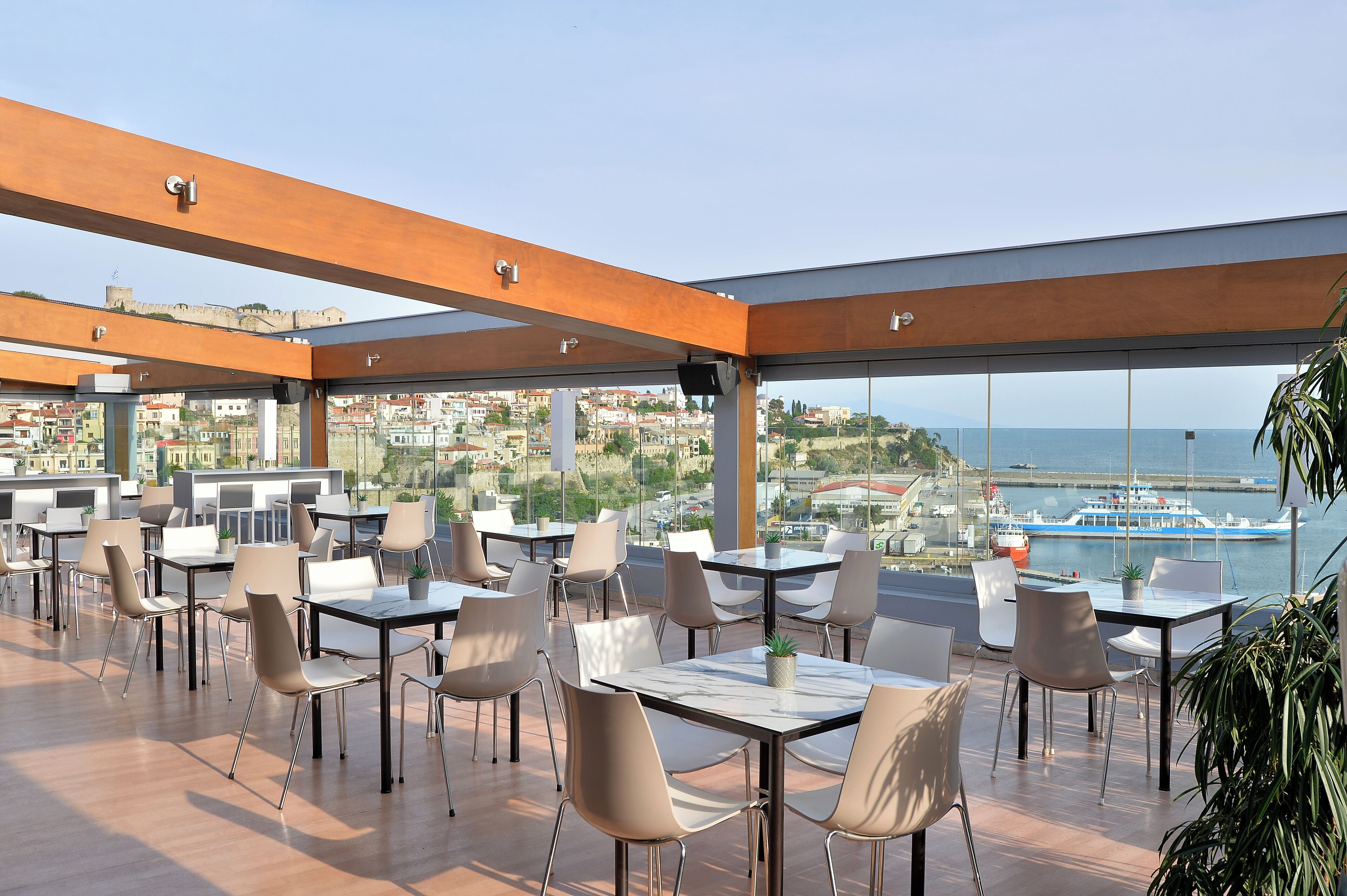 A rooftop dining area with tables and chairs, large windows offering a view of a marina and cityscape.