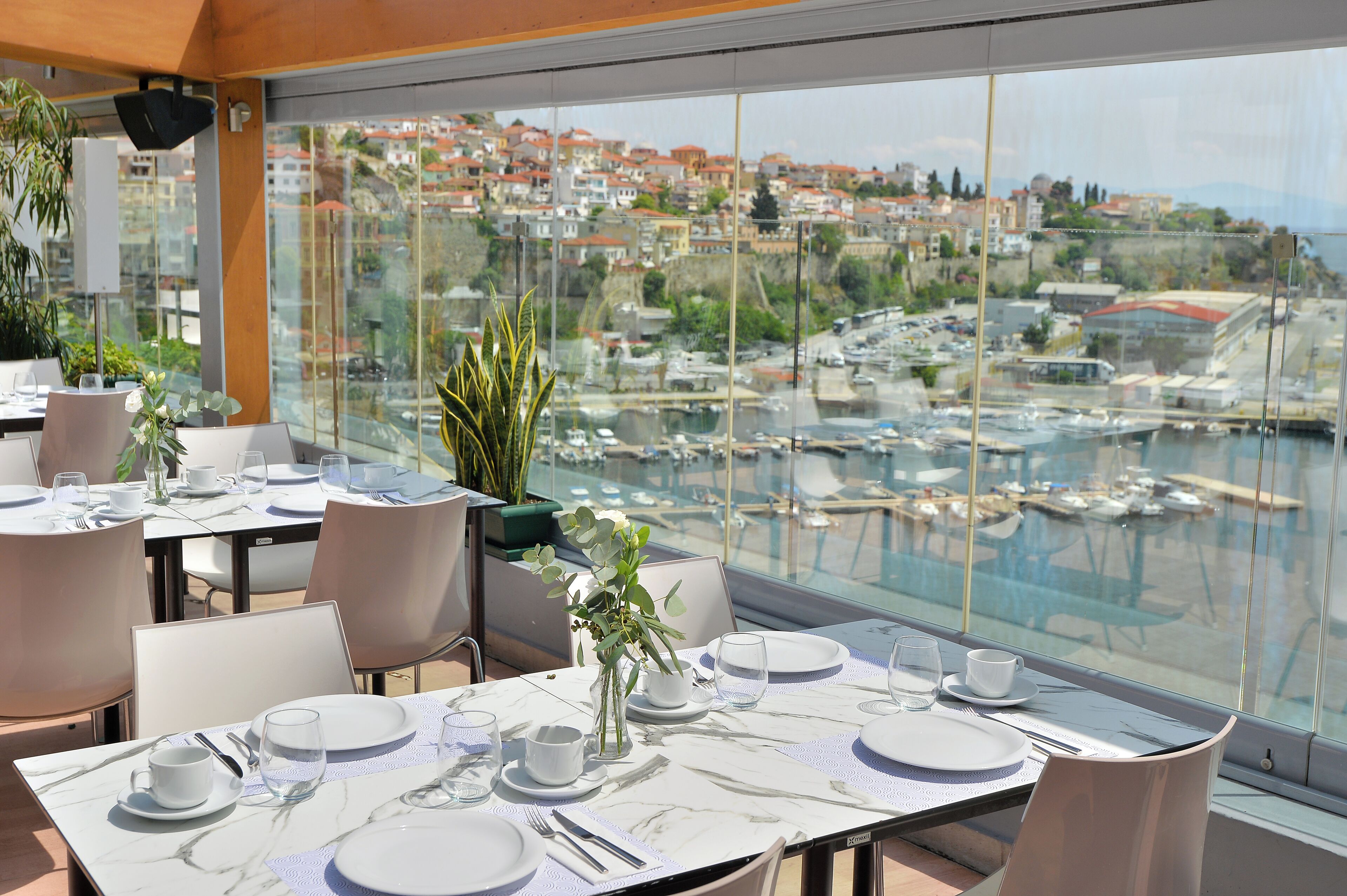 A dining area with tables set for two, large windows offering a view of a marina and town.