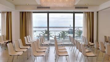 Uma sala de conferências com fileiras de cadeiras brancas e amplas janelas que oferecem vista para o mar e um horizonte urbano ao longe.
