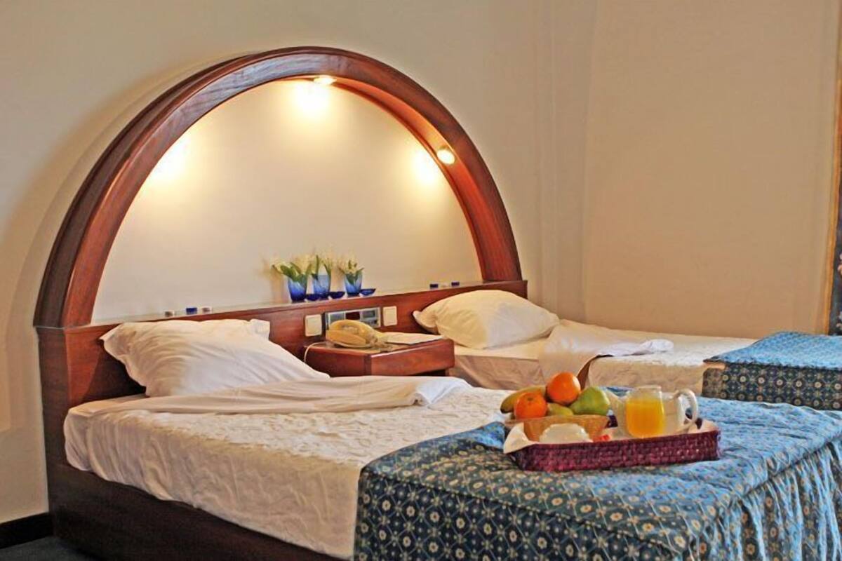 Quarto de hotel com duas camas, uma cabeceira de madeira, uma mesa de cabeceira com abajur e uma cesta de frutas na cama.