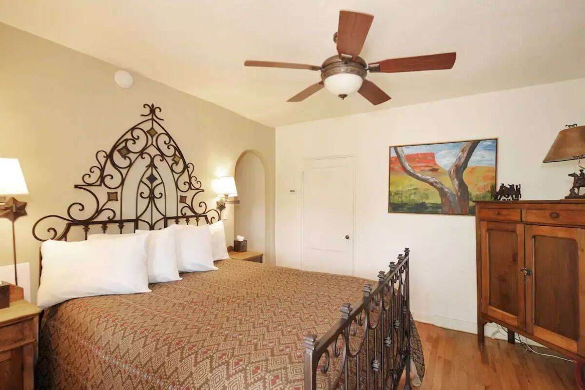 A bedroom with a patterned bedspread, a decorative headboard, a ceiling fan, a painting on the wall, and a wooden cabinet.