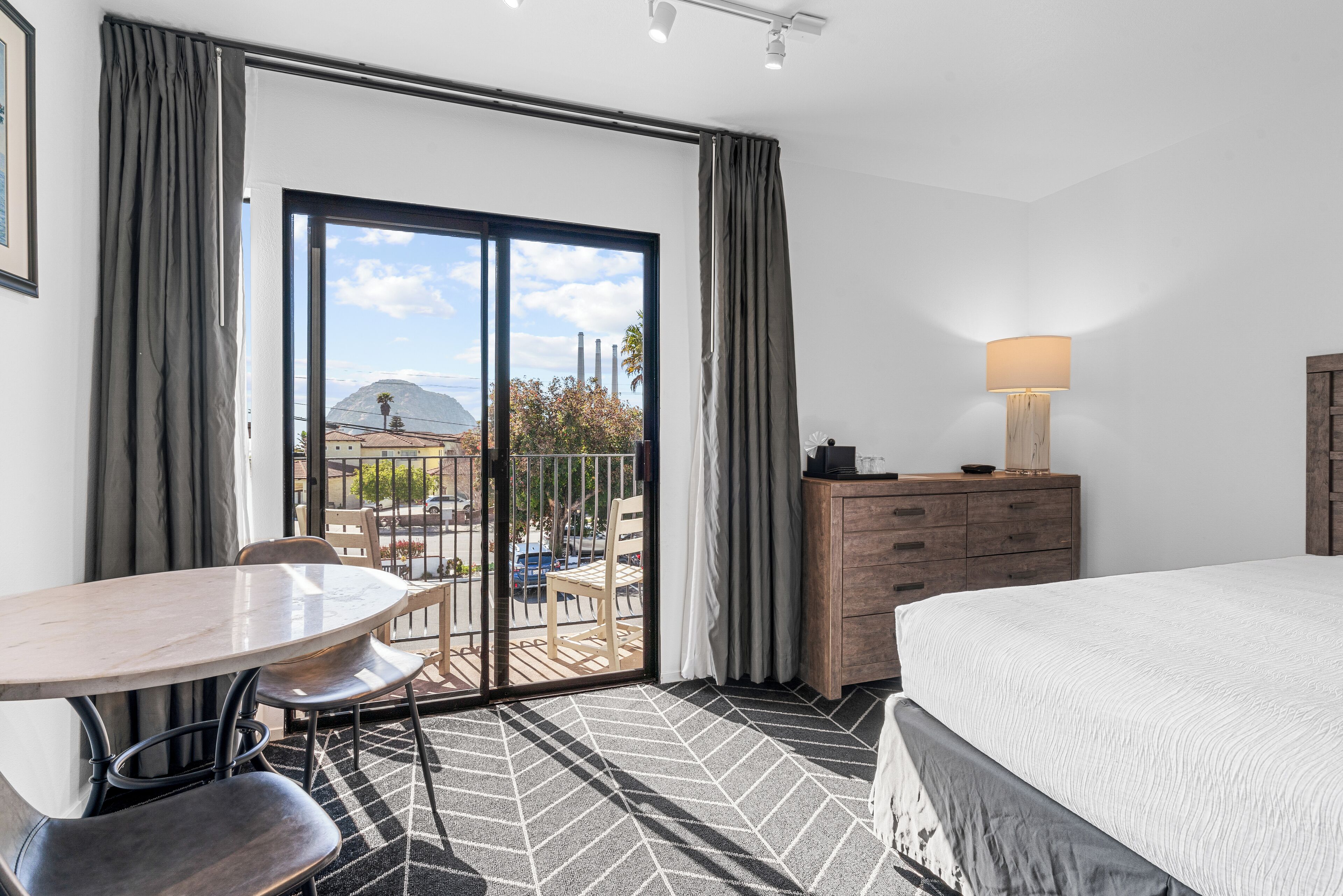 Una camera d'albergo con un letto, una cassettiera in legno, un tavolo con sedie e un balcone con vista sulla città e sulle montagne.