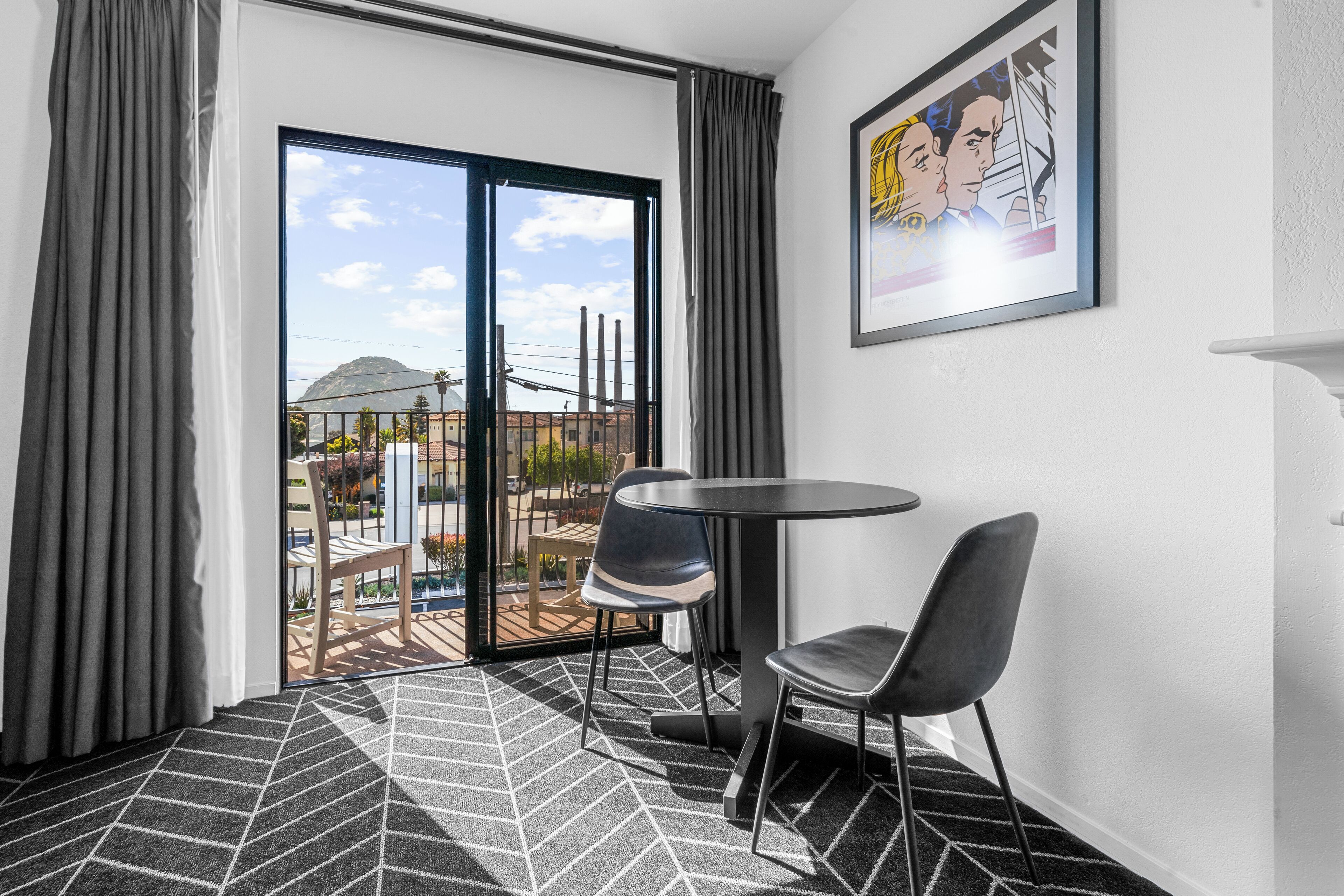 A hotel room with a balcony, a round table, two chairs, a patterned rug, and a framed picture on the wall.