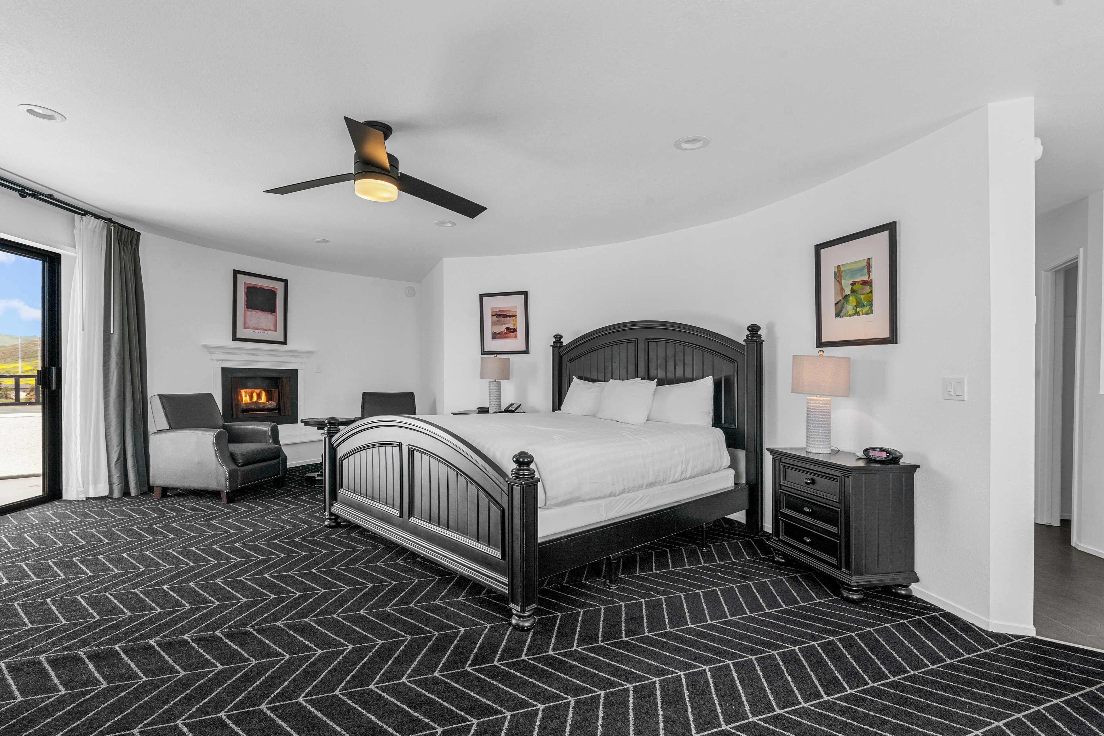 A modern bedroom with a large bed, a ceiling fan, a fireplace, and a view of the outdoors.