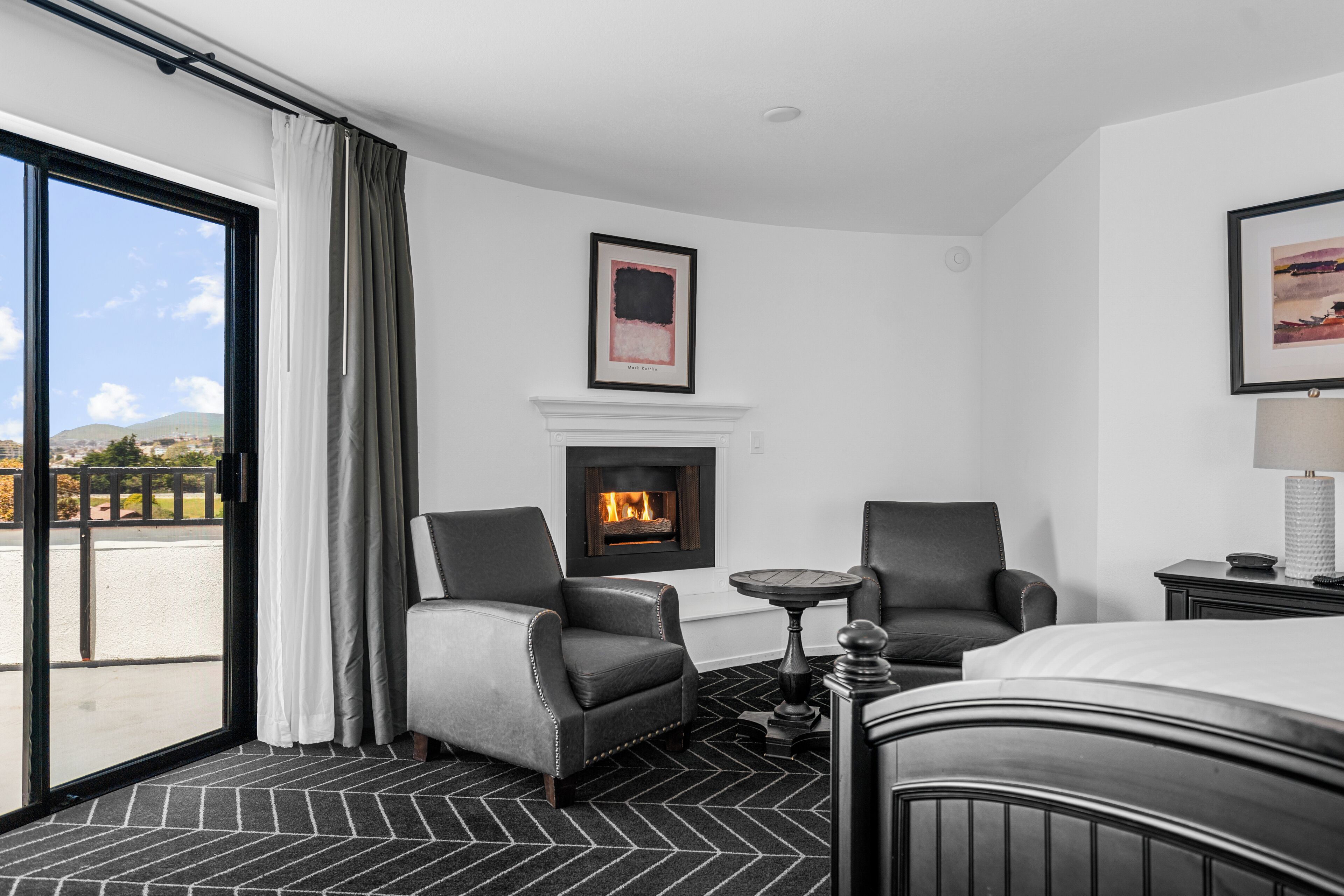 A hotel room with a fireplace, two armchairs, a bed, and a view of the outdoors.