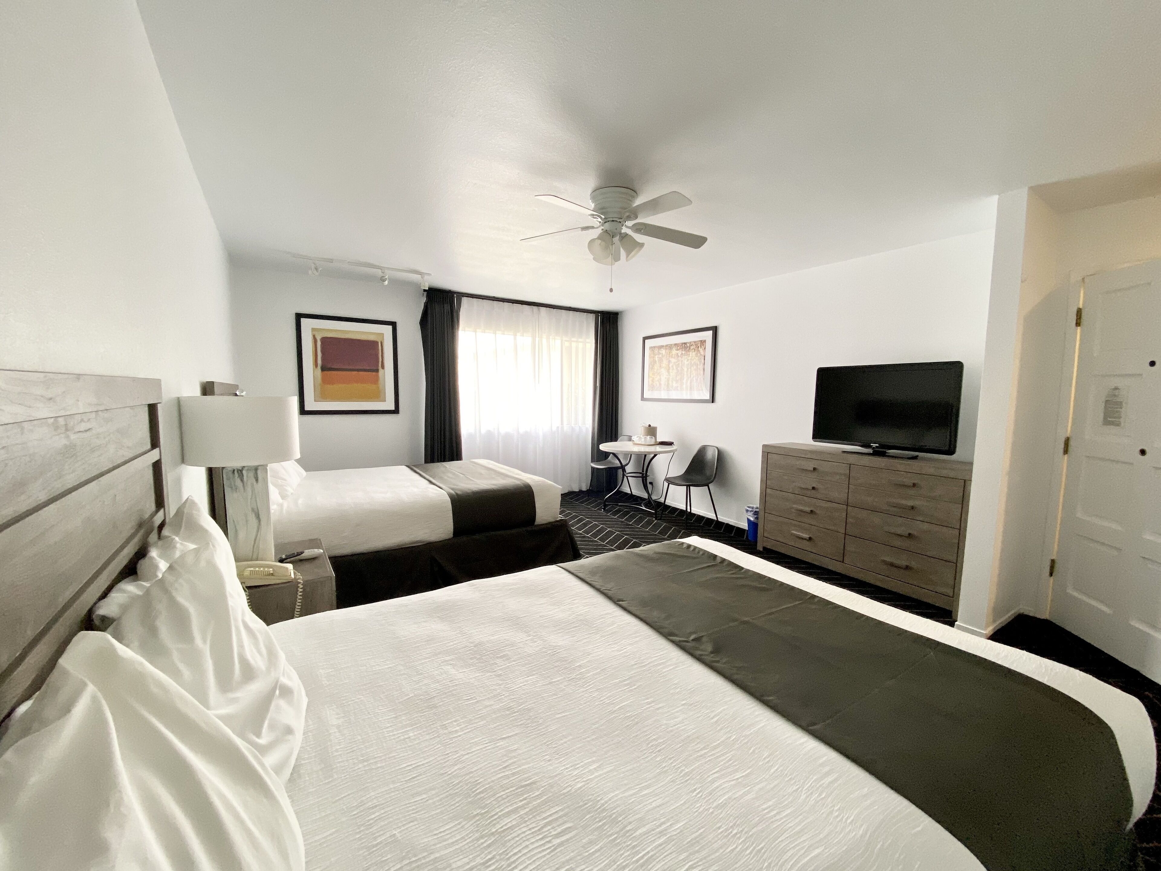 A hotel room with a large bed, a desk with a chair, a television, a ceiling fan, and artwork on the walls.