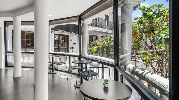 Un espacio interior moderno con balcón, que cuenta con una mesa redonda y sillas, ventanas amplias y vistas a un edificio con balcones.