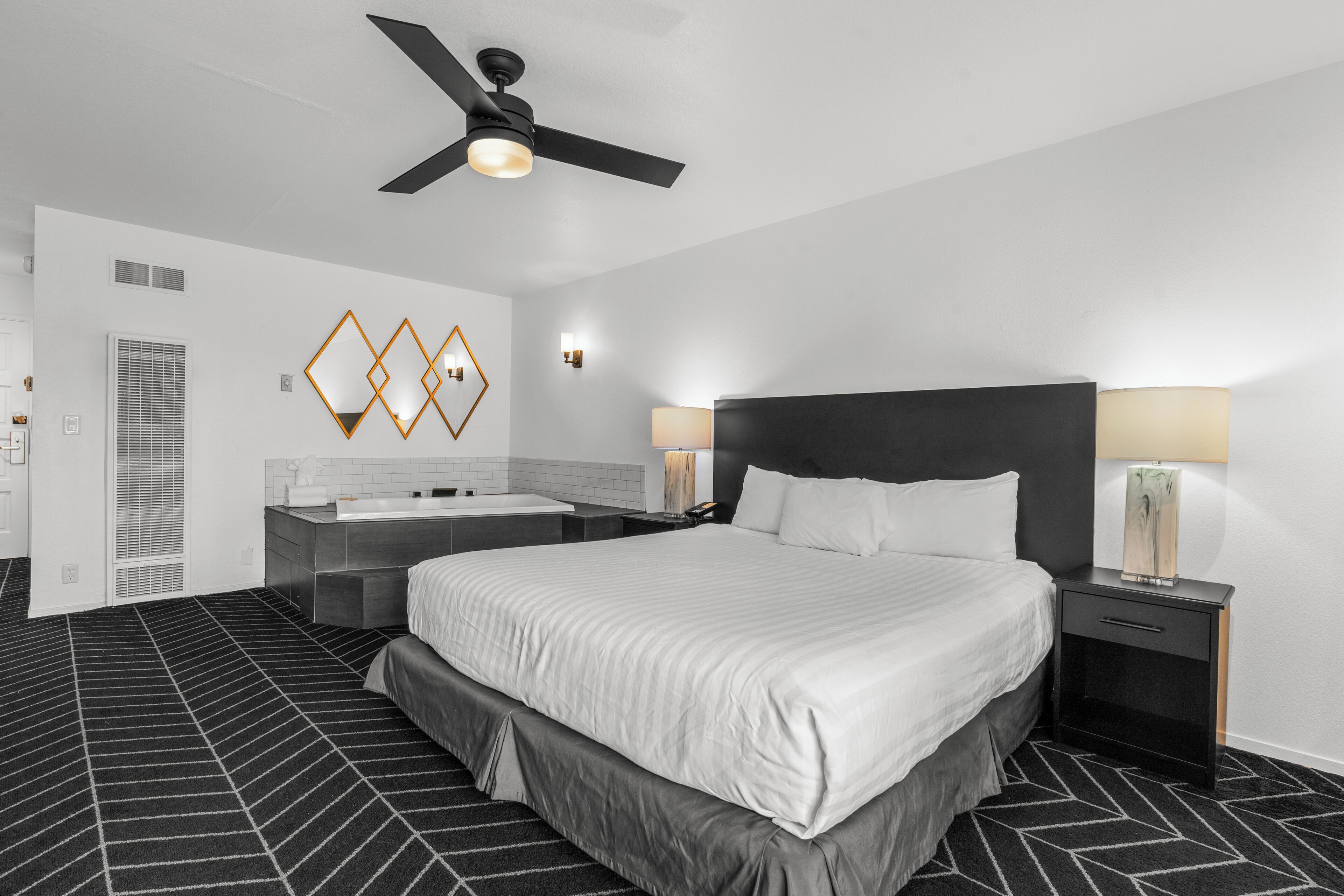 A modern hotel room with a large bed, a bedside table with a lamp, a ceiling fan, and a decorative wall piece.