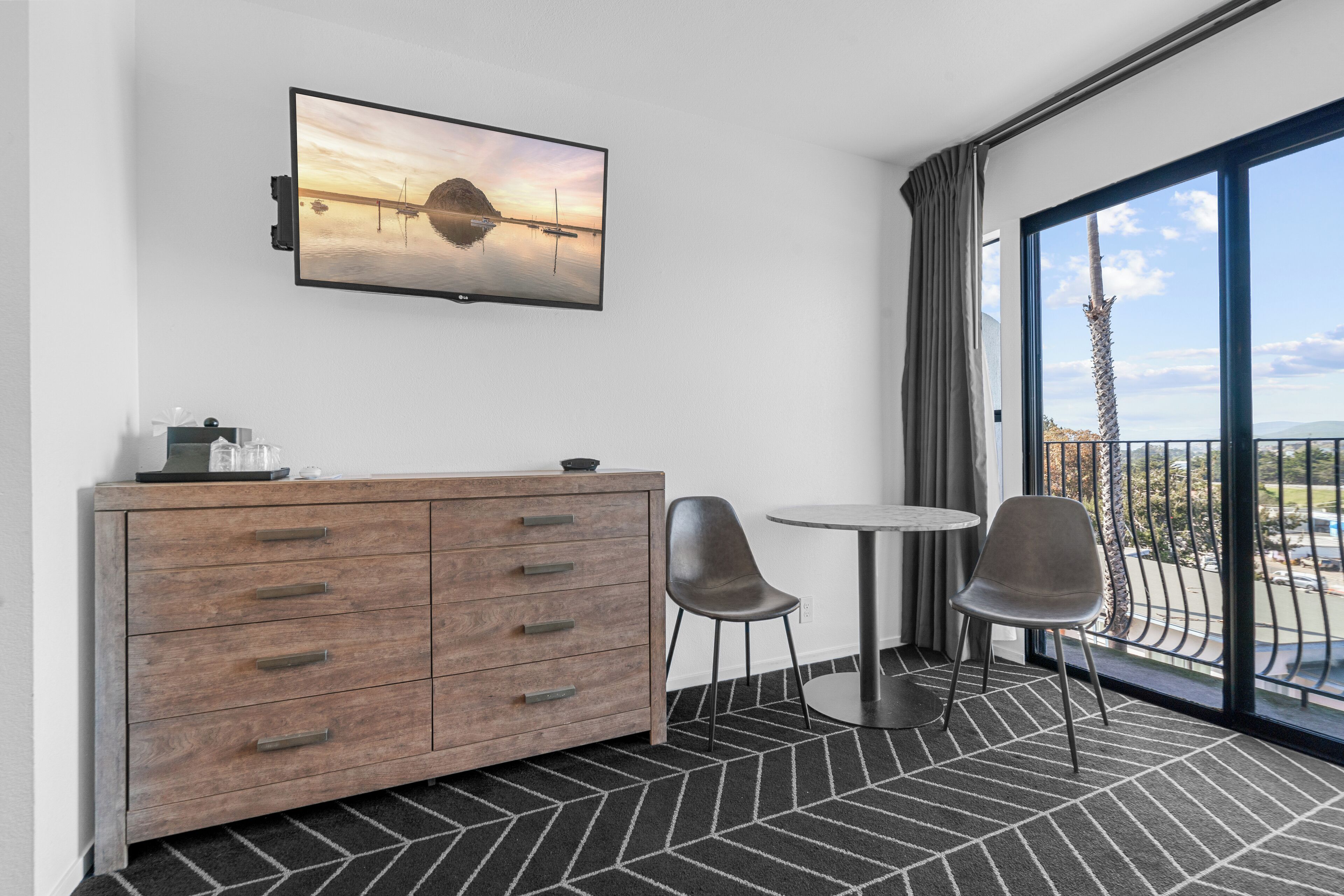 A hotel room with a large window, a wooden dresser, two chairs, and a flat-screen TV mounted on the wall.