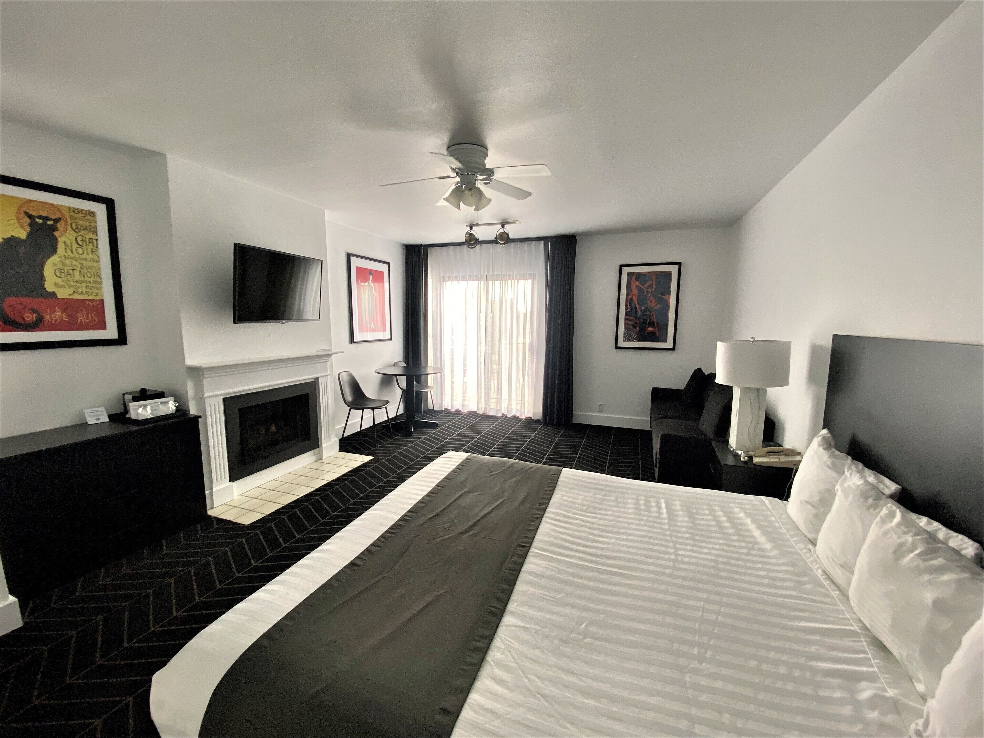 A hotel room with a large bed, a flat-screen TV, a fireplace, a ceiling fan, and artwork on the walls.