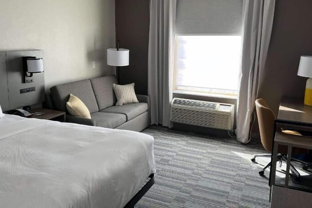 A hotel room with a bed, a sofa, a desk, a chair, a lamp, and a window with curtains.