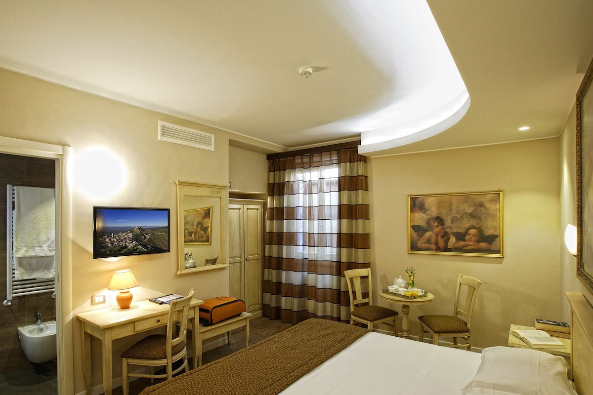 A hotel room with a bed, desk, chair, and a small table with a vase.