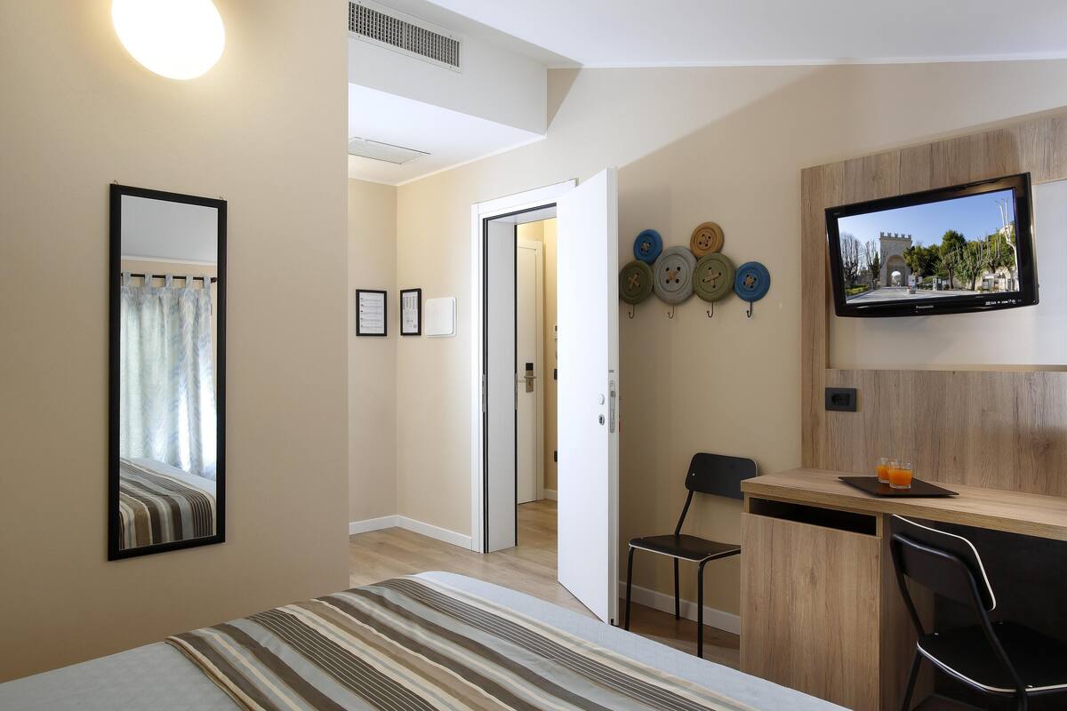 A hotel room with a bed, a desk with a chair, a TV, and a wall-mounted clock.