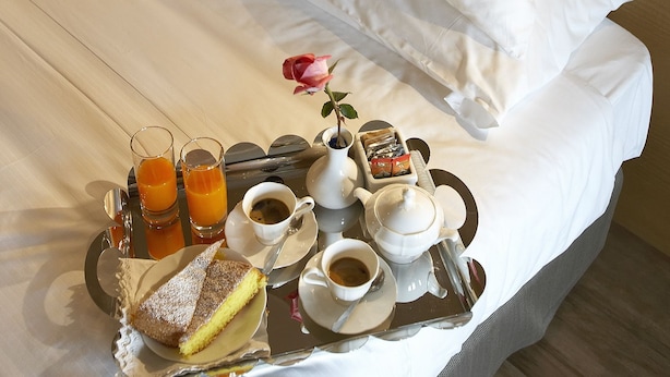 Una cama bien hecha con sábanas y almohadas blancas, una bandeja con alimentos para el desayuno, incluyendo una porción de pastel, dos vasos de zumo de naranja y un jarrón con una rosa rosa.