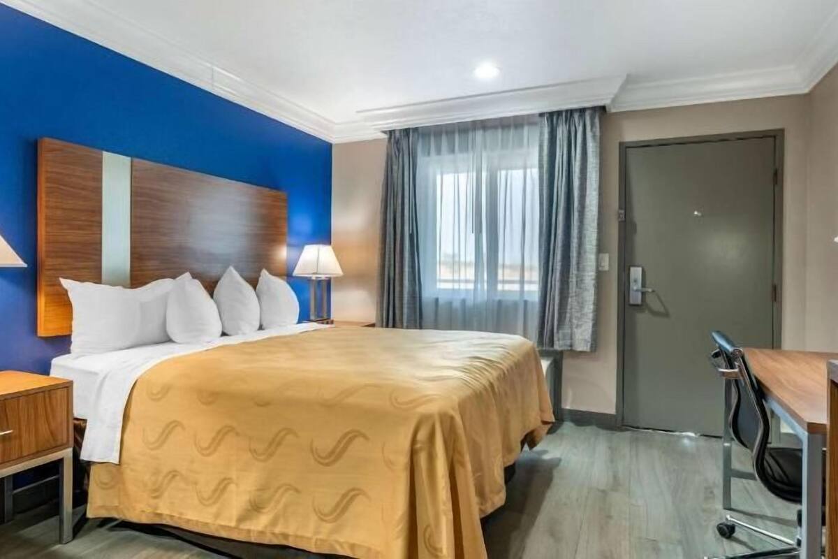 A hotel room with a bed, two bedside tables, a desk with a chair, a window with curtains, and a door.