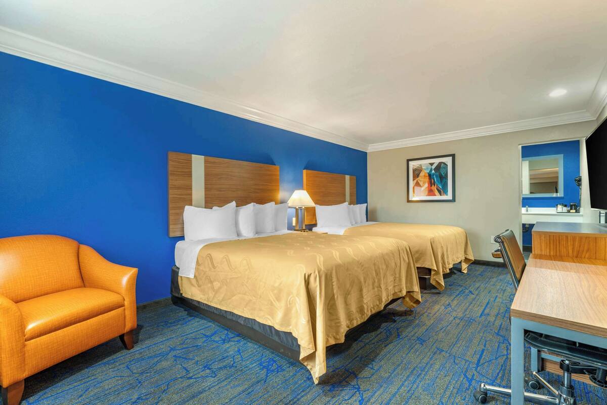 A hotel room with two beds, a desk, a chair, and a bathroom mirror.