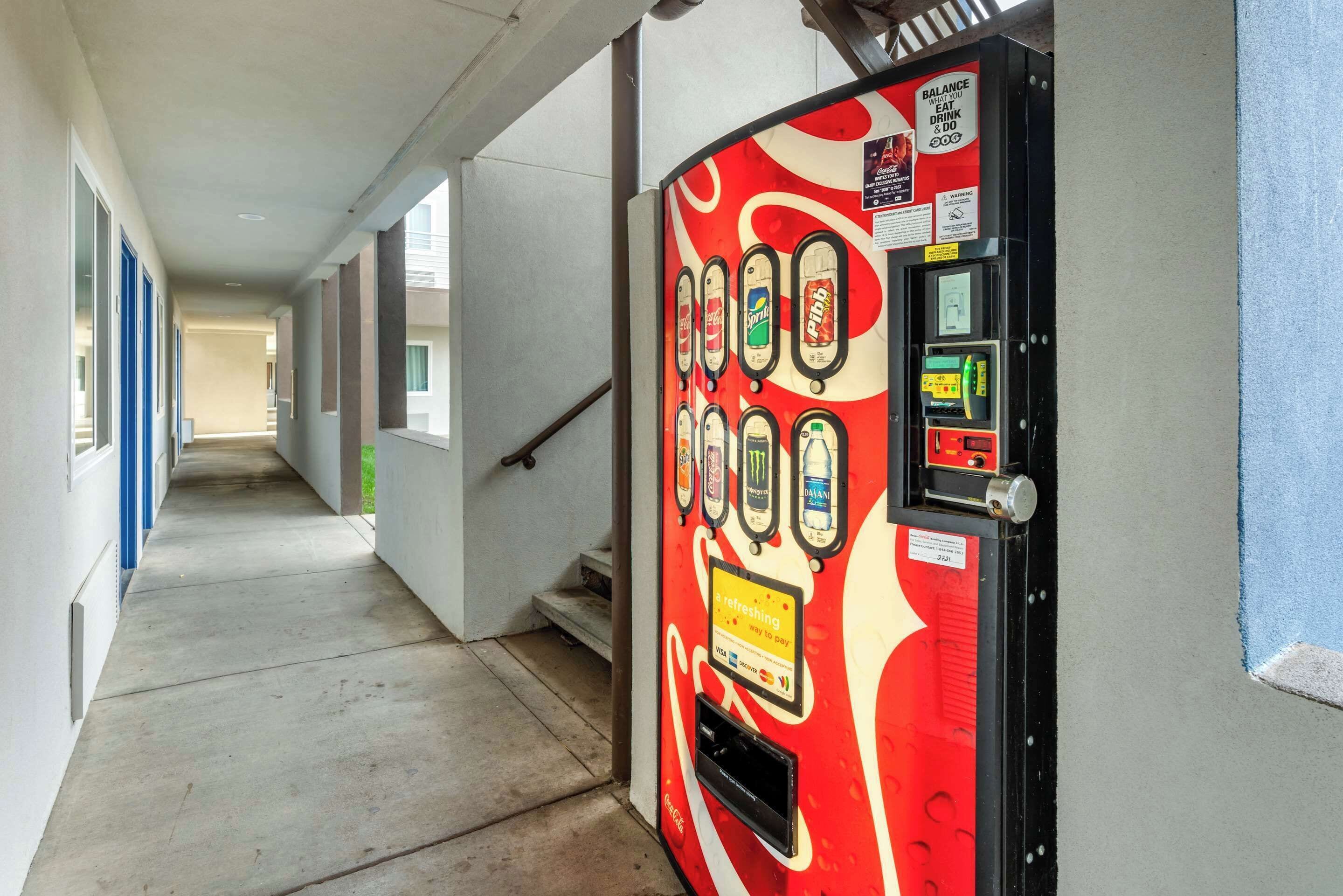 A vending machine with various drink options in a hallway with a concrete floor and blue-framed windows.