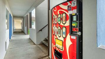 A vending machine with various drink options in a hallway with a concrete floor and blue-framed windows.