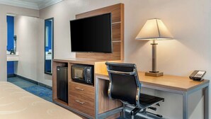 A hotel room with a flat-screen TV mounted on the wall, a desk with a lamp, a microwave, and a chair.