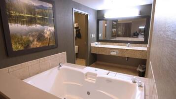 A modern bathroom with a large bathtub, a granite countertop vanity, a large mirror, and a framed landscape painting on the wall.