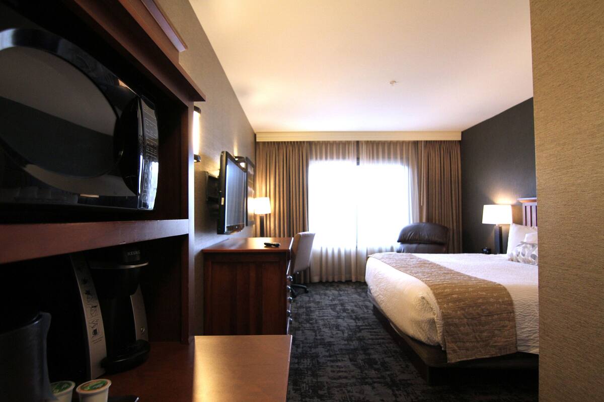 A hotel room with a bed, a desk with a coffee maker, a TV, and a window with curtains.