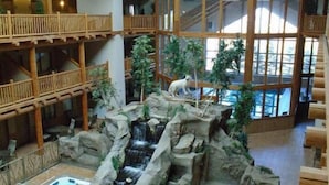 A multi-level indoor space with wooden architecture, a central rock formation with a waterfall, and two hot tubs.